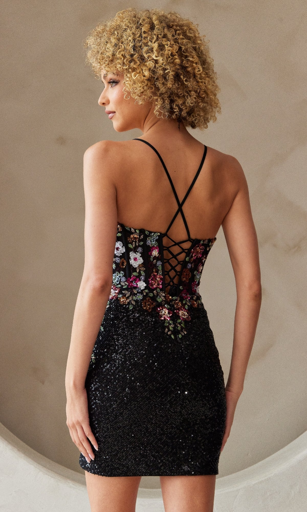 A woman stands with her back to the camera, wearing the Floral-Embroidered Short Sequin Hoco Dress 7054S, featuring a black sequin skirt, corset-style floral embroidered bodice, and crisscross straps.